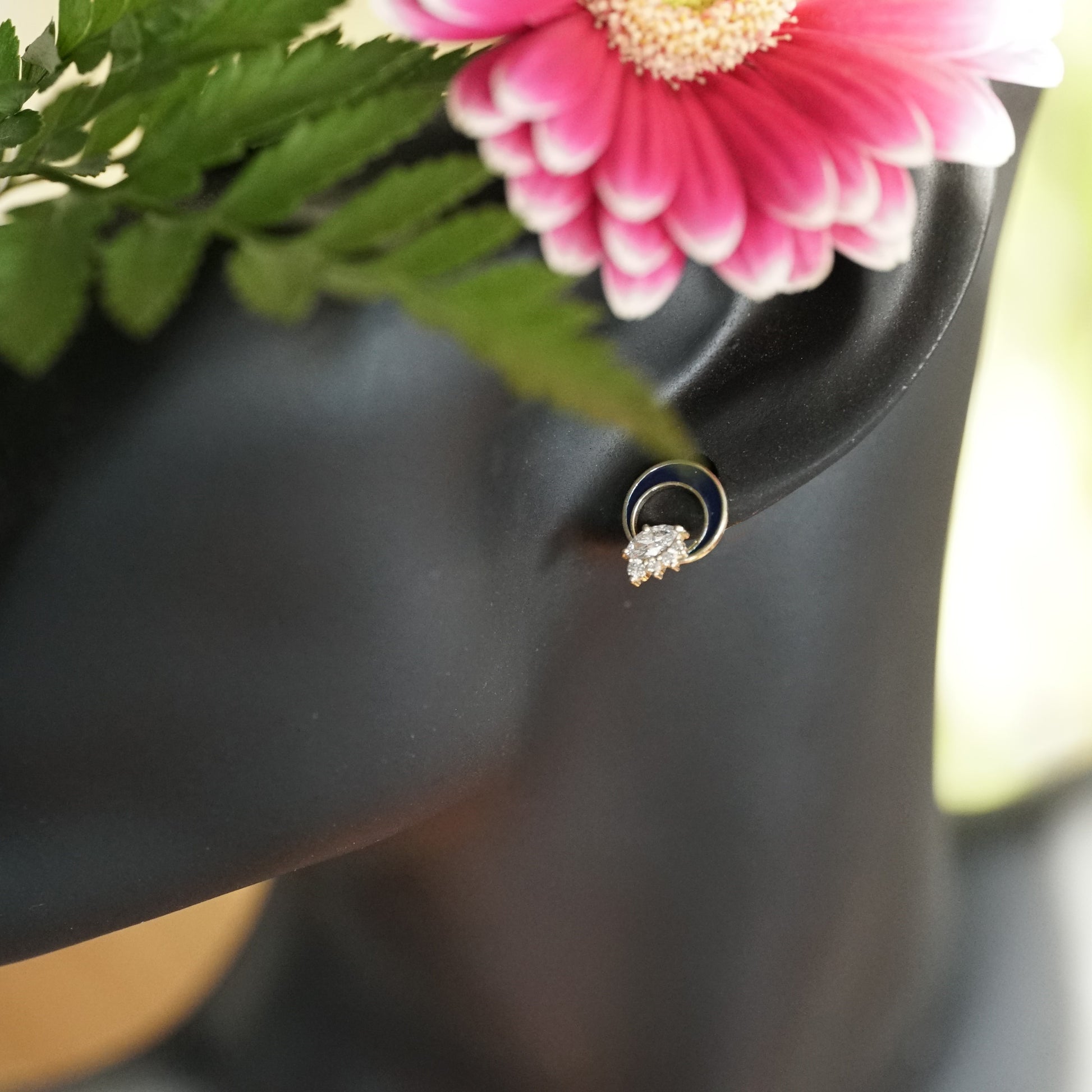 a pair of diamond button earrings with one large marquise and 4 small round diamonds at the base of a crescent shape element filled with blue enamel displayed on a manikin for scale with pink flowers and green foliage 