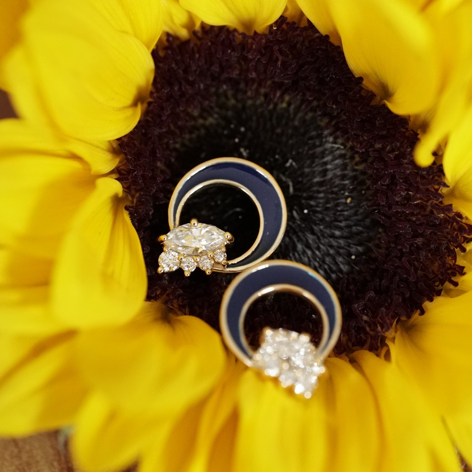 a pair of diamond button earrings with one large marquise and 4 small round diamonds at the base of a crescent shape element filled with blue enamel resting in the middle of a vibrant yellow sunflower