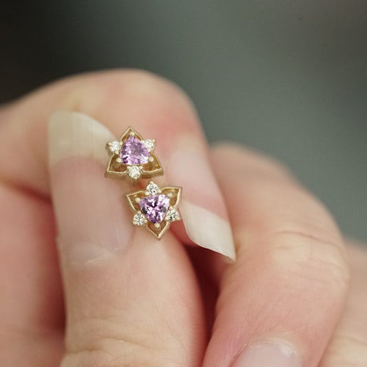 a pair of yellow gold triangular shaped studs featuring a pink sapphire trillion center stone and 3 round diamond accents held in a light skinned left hand