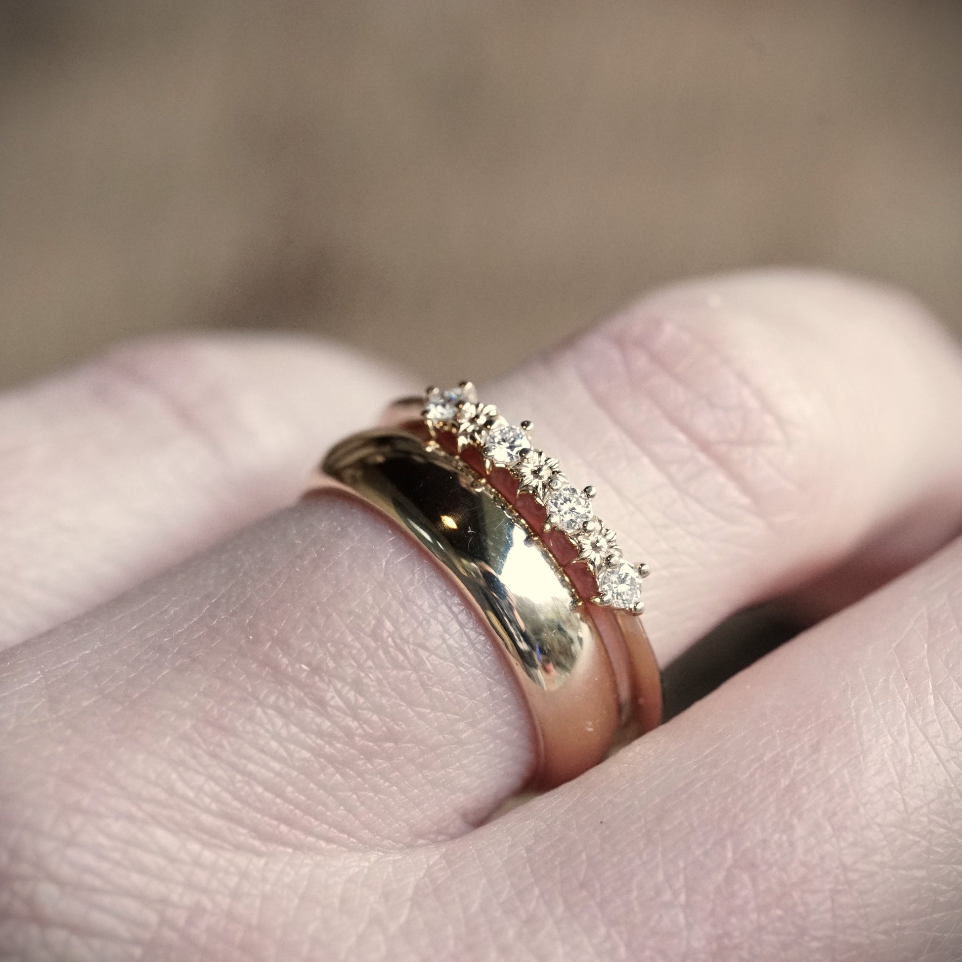 a yellow gold ring with 3 flower elements and 4 diamonds next to a wide yellow gold band on a light skinned left hand