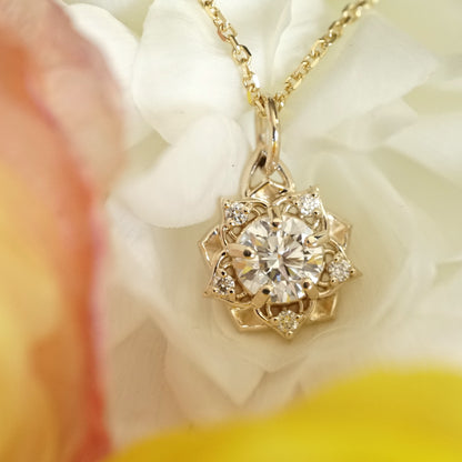 a yellow gold pendant with a large diamond center stone and 5 diamond accents in a floral design on an open link chain rests on a white flower with yellow and orange flowers in the foreground