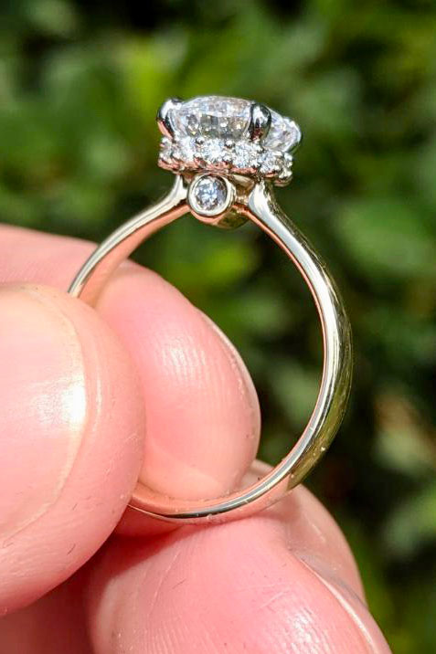 a yellow gold and diamond engagement ring is held in a light skin tone hand in front of green shrubs