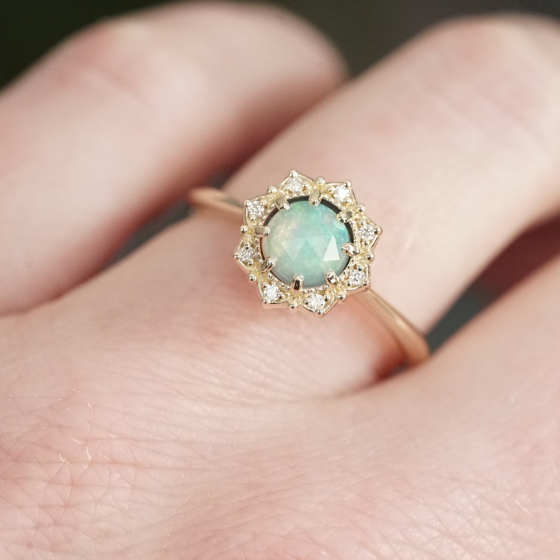 a yellow gold ring featuring a rose cut opal center stone in an 8 prong setting and with a floral diamond halo on a light skinned left hand