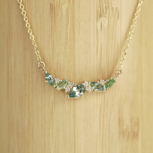 a yellow gold pendant features green and teal gemstones paired with round white diamonds in a playful design hangs on an open link chain in front of a bamboo background