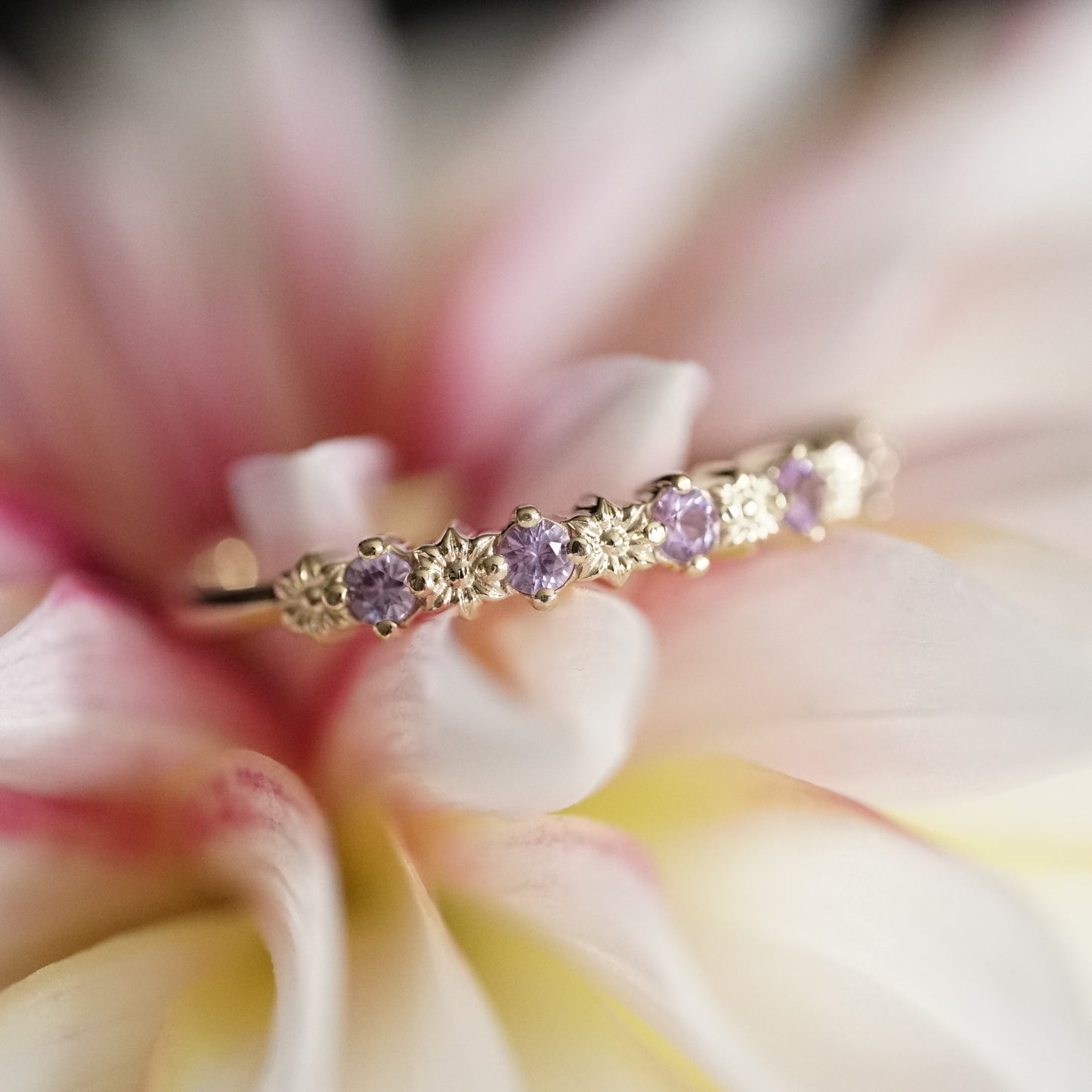 a yellow gold ring with 6 carved flowers and 5 light purple sapphires rests on a pink and yellow flower