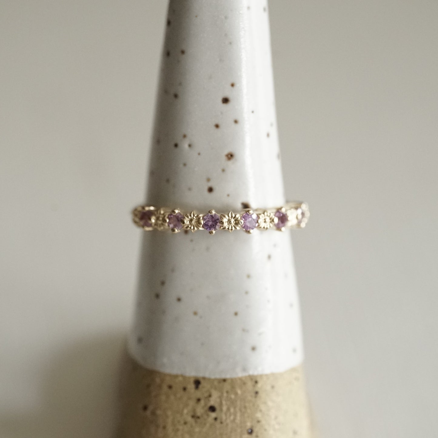 a yellow gold ring with 6 carved flowers and 5 light purple sapphires on a white ceramic display