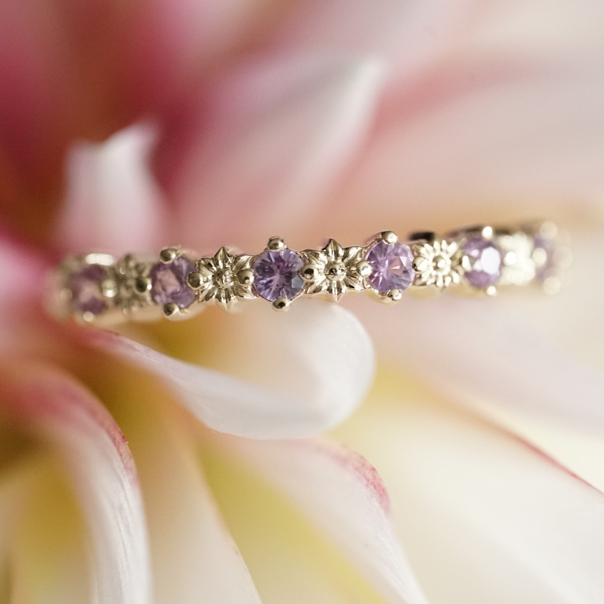 a macro photo of a a yellow gold ring with 6 carved flowers and 5 light purple sapphires on a pink and white flower