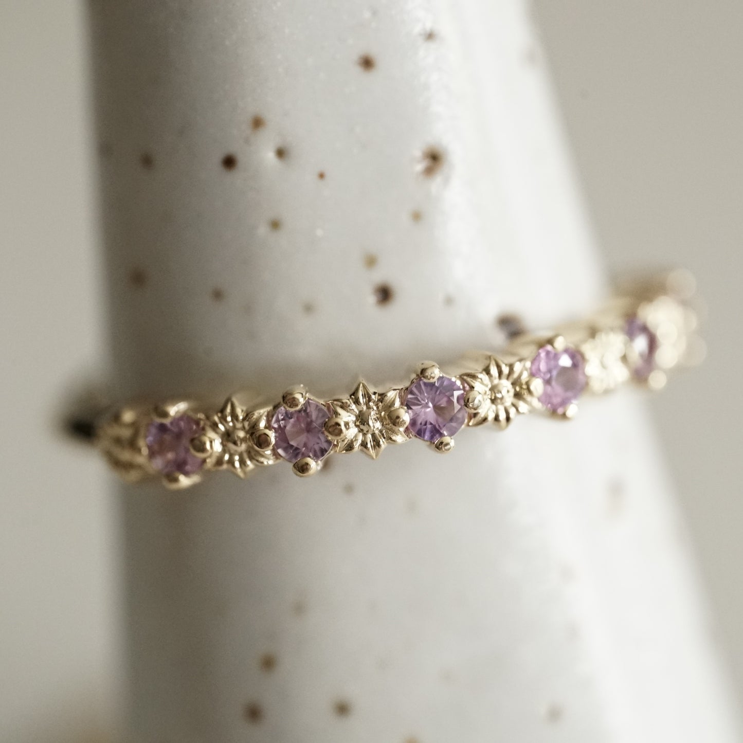 a yellow gold ring with 6 carved flowers and 5 light purple sapphires on a white ceramic display