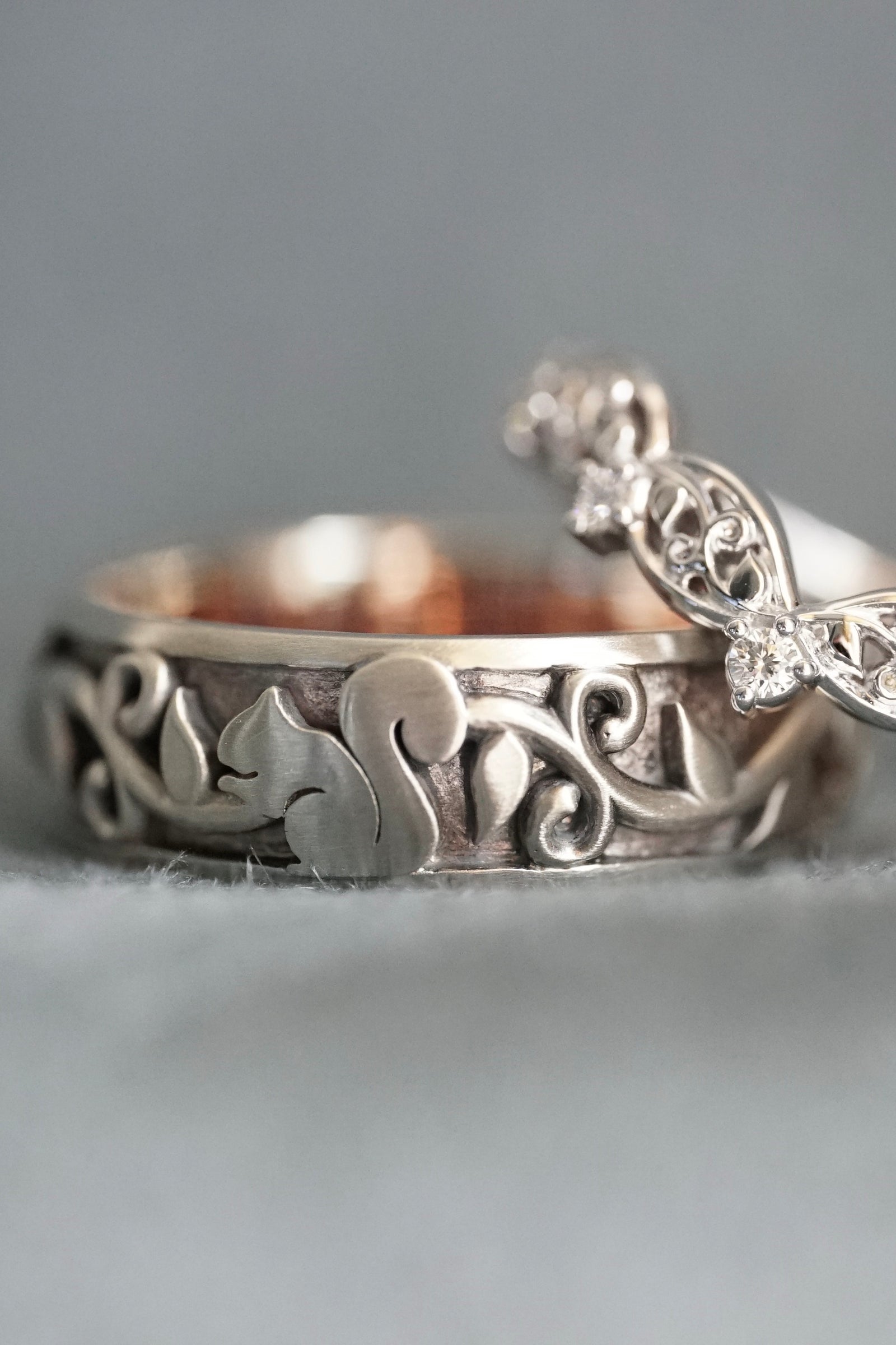 a white gold wedding band with a squirrel next to a white gold and diamond wedding band on a light grey fabric base