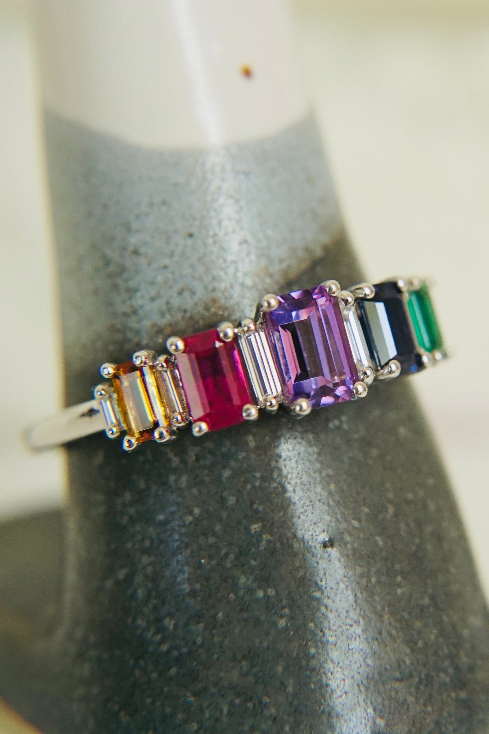 a white gold ring with mutli colored emerald gemstones on a ceramic ring display