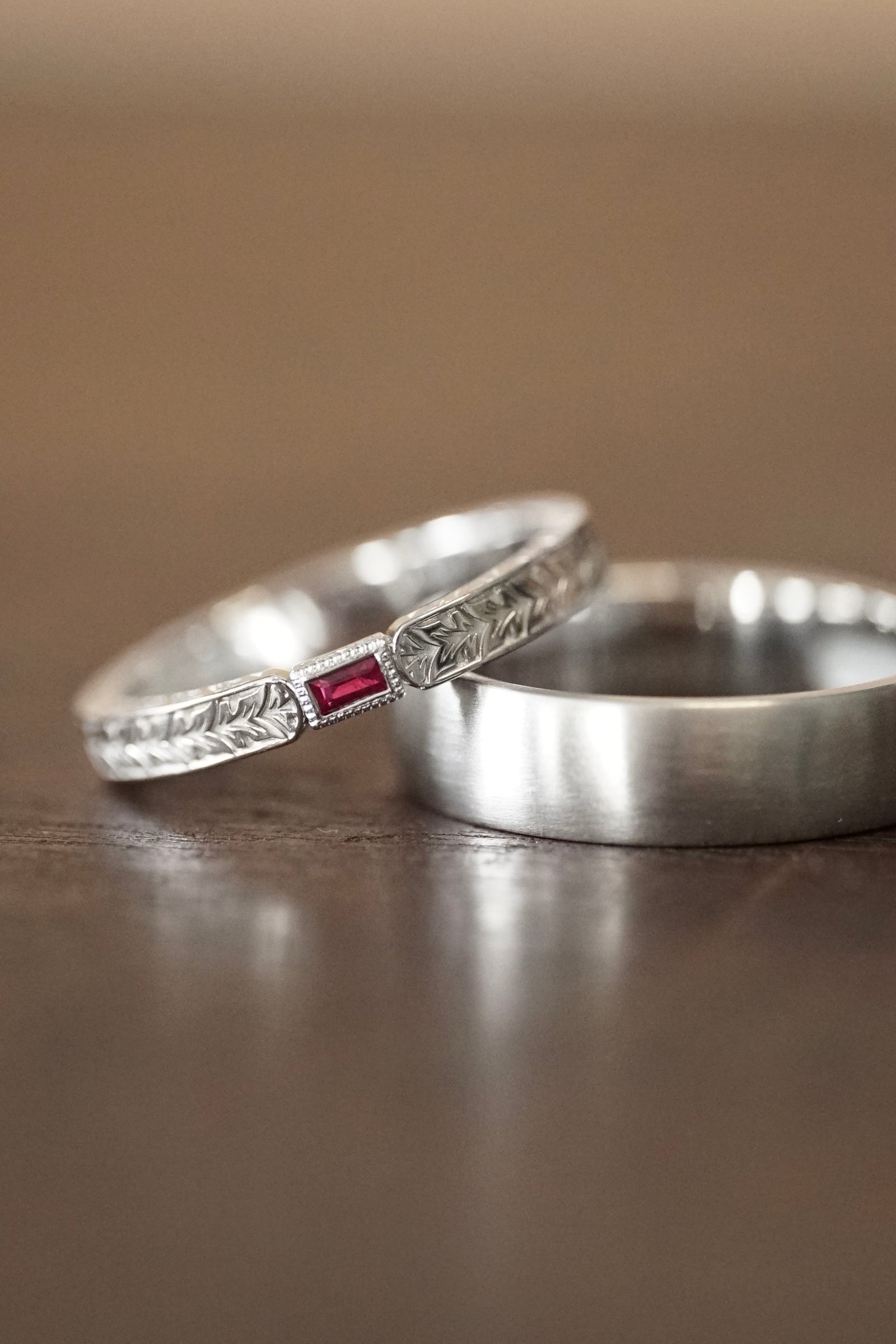 an engraved white gold and ruby band rests on a white gold gents band on a wood base