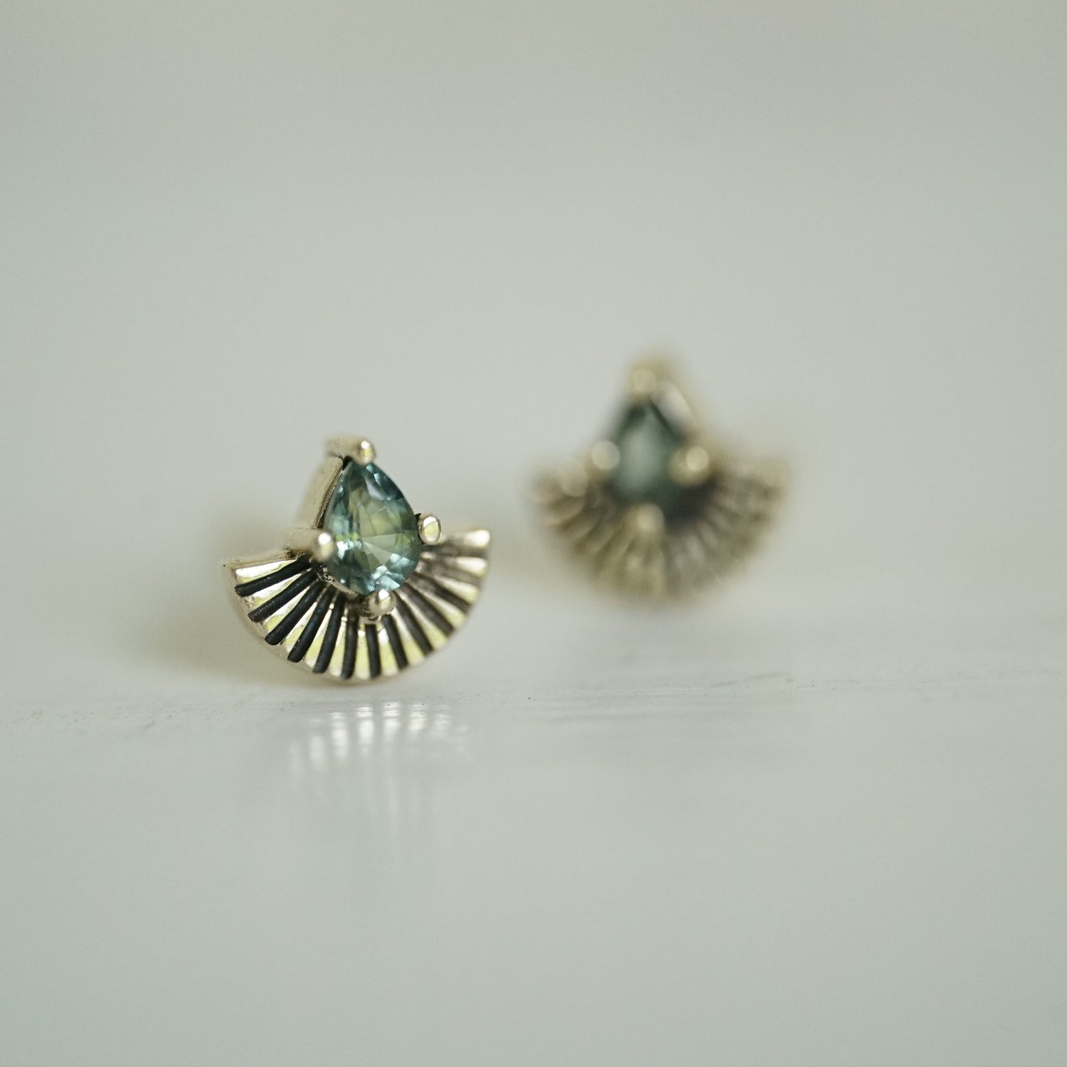 a pair of yellow gold earrings with pear shape green sapphires and fan shapes underneath on a white background