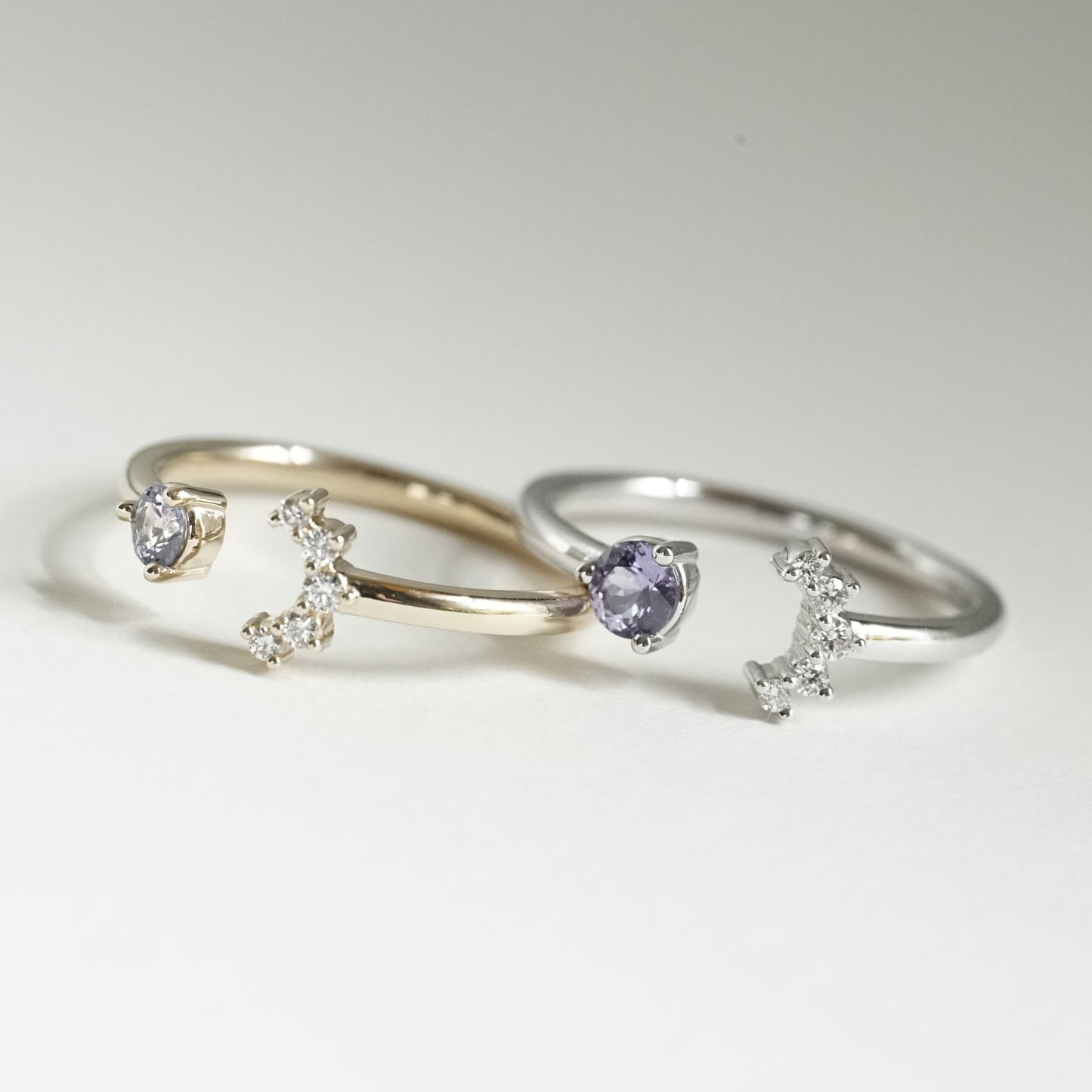 a white gold open style stacking ring with a light blue sapphire on the left and 5 diamonds on the right in a crescent shape next to a yellow gold open style stacking ring with a light blue sapphire on the left and 5 diamonds on the right in a crescent shape on a white background