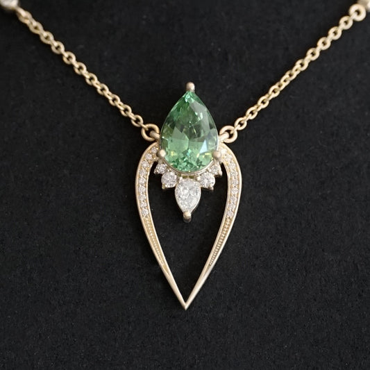 a yellow gold pendant with a vibrant green tourmaline pear shape and diamond accents suspended on a cable chain and laying on a matte black background