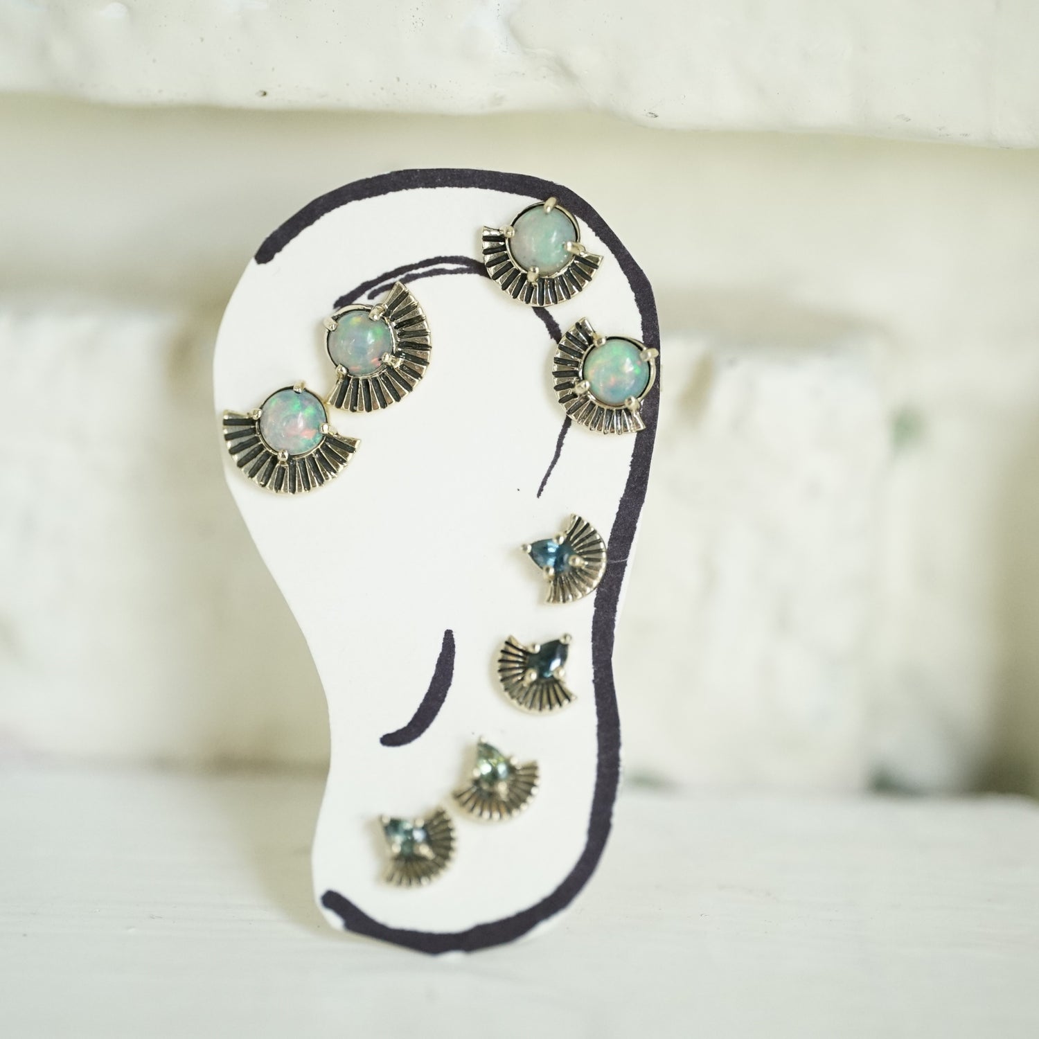 A hand drawn ear with 4 pairs of art deco inspired stud earrings, 2 pairs featuring opals and 2 pairs featuring pear shape sapphires