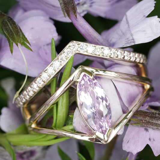 a macro photo of a yellow gold ring with a marquise cut lavender spinel between two V shape shadow style bands next to a V shape pave set diamond wedding band shown in purple and green flowers