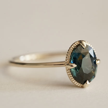 a macro photo of an oval teal sapphire is set in a yellow gold ring with a beaded detail behind the stone on a white matte background