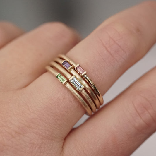 A set of 4 yellow gold stacking bands with various metal texture finishes and multiple colors of gems on a light-skinned left hand