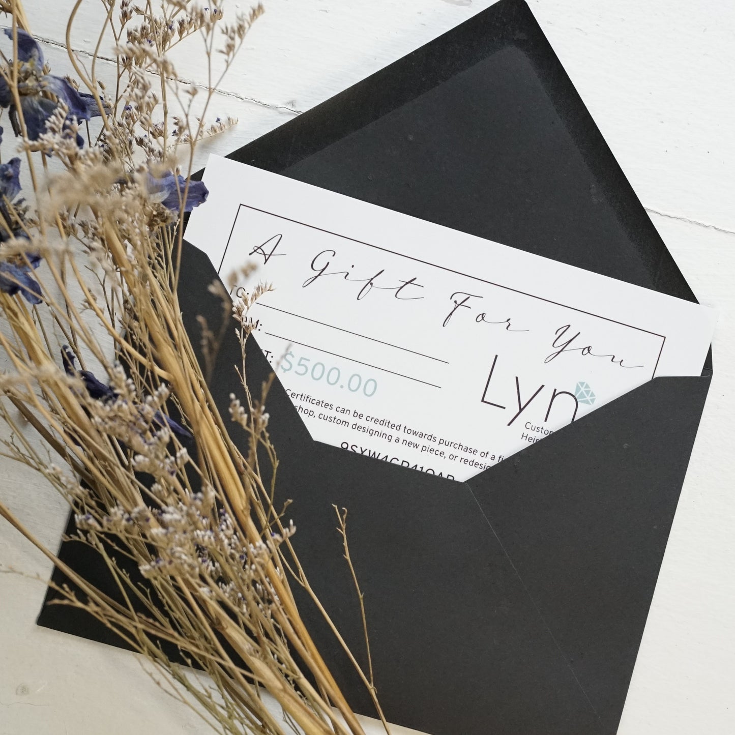 a white gift certificate shown halfway out of a black envelope with a bouquet of dried flowers on top and on a white wood backdrop