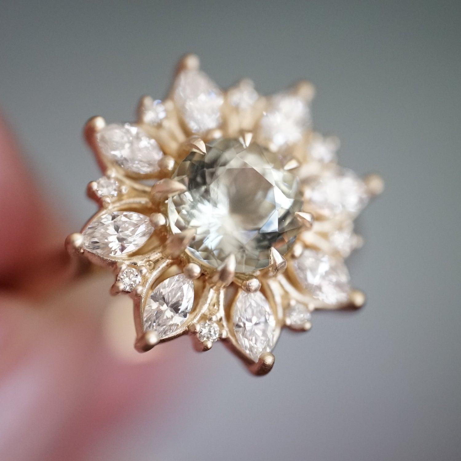 a macro photo of a yellow gold ring with a very light green center stone and mixed shape diamond halo
