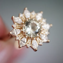 a macro photo of a yellow gold ring with a very light green center stone and mixed shape diamond halo