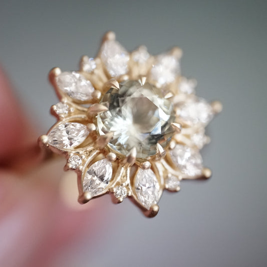 a macro photo of a yellow gold ring with a very light green center stone and mixed shape diamond halo