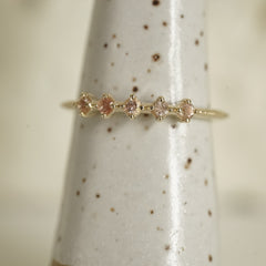 a yellow gold ring with 5 round light pink Oregon sunstones and migraine detail on the shank on a white ceramic display