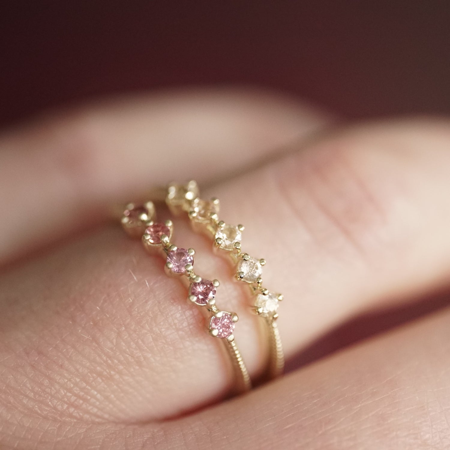 a yellow gold ring with 5 round light pink Oregon sunstones is stacked next to a yellow gold ring with 5 pink padparadscha sapphires on a light skinned left hand