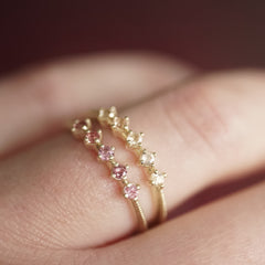 a yellow gold ring with 5 round light pink Oregon sunstones is stacked next to a yellow gold ring with 5 pink padparadscha sapphires on a light skinned left hand