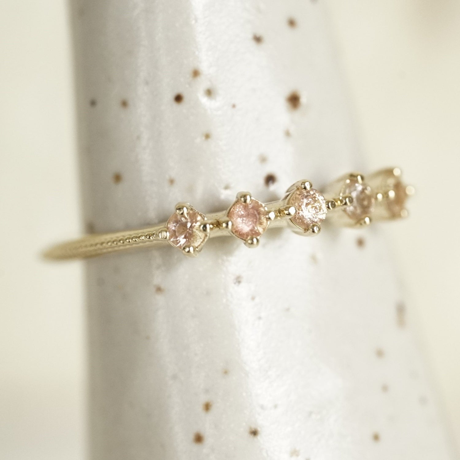 a yellow gold ring with 5 round light pink Oregon sunstones and migraine detail on the shank on a white ceramic display