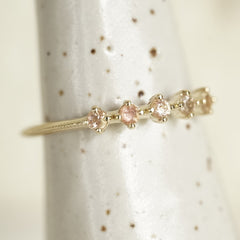 a yellow gold ring with 5 round light pink Oregon sunstones and migraine detail on the shank on a white ceramic display