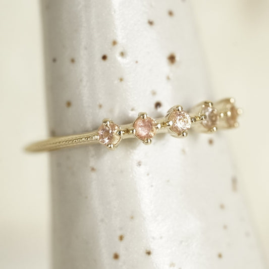 a yellow gold ring with 5 round light pink Oregon sunstones and migraine detail on the shank on a white ceramic display