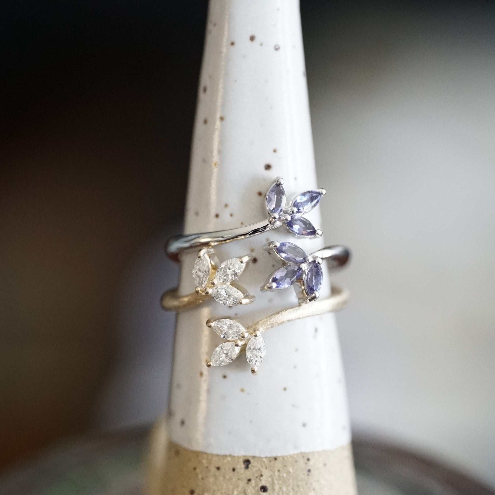 A polished white gold and purple iolite leaf inspired ring stacked on top of a matte yellow gold and diamond leaf inspired ring on a white ceramic display