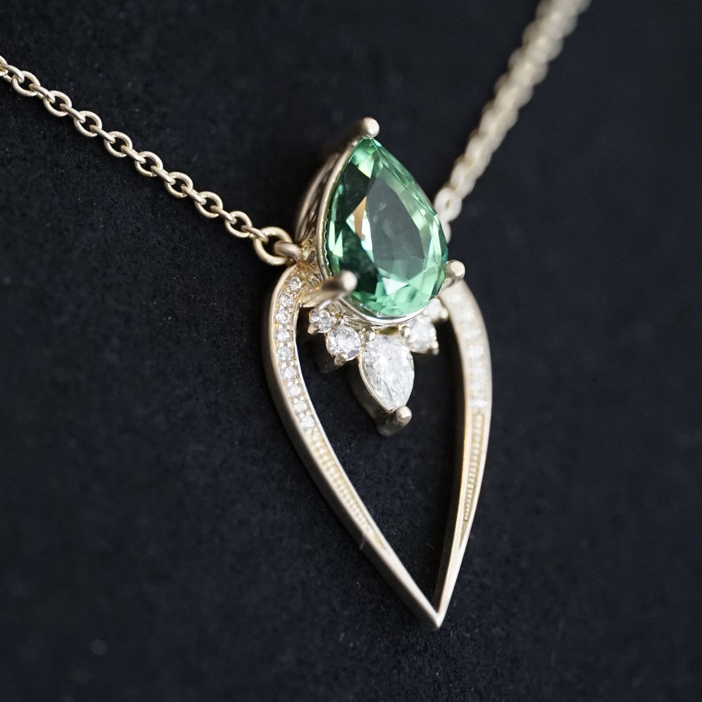 a yellow gold pendant with a vibrant green tourmaline pear shape and diamond accents suspended on a cable chain and laying on a matte black background