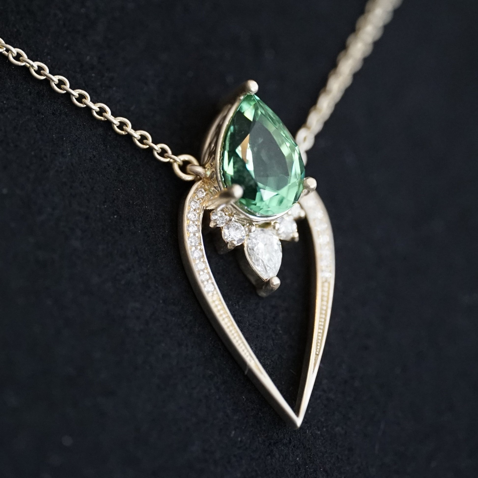 a yellow gold pendant with a vibrant green tourmaline pear shape and diamond accents suspended on a cable chain and laying on a matte black background