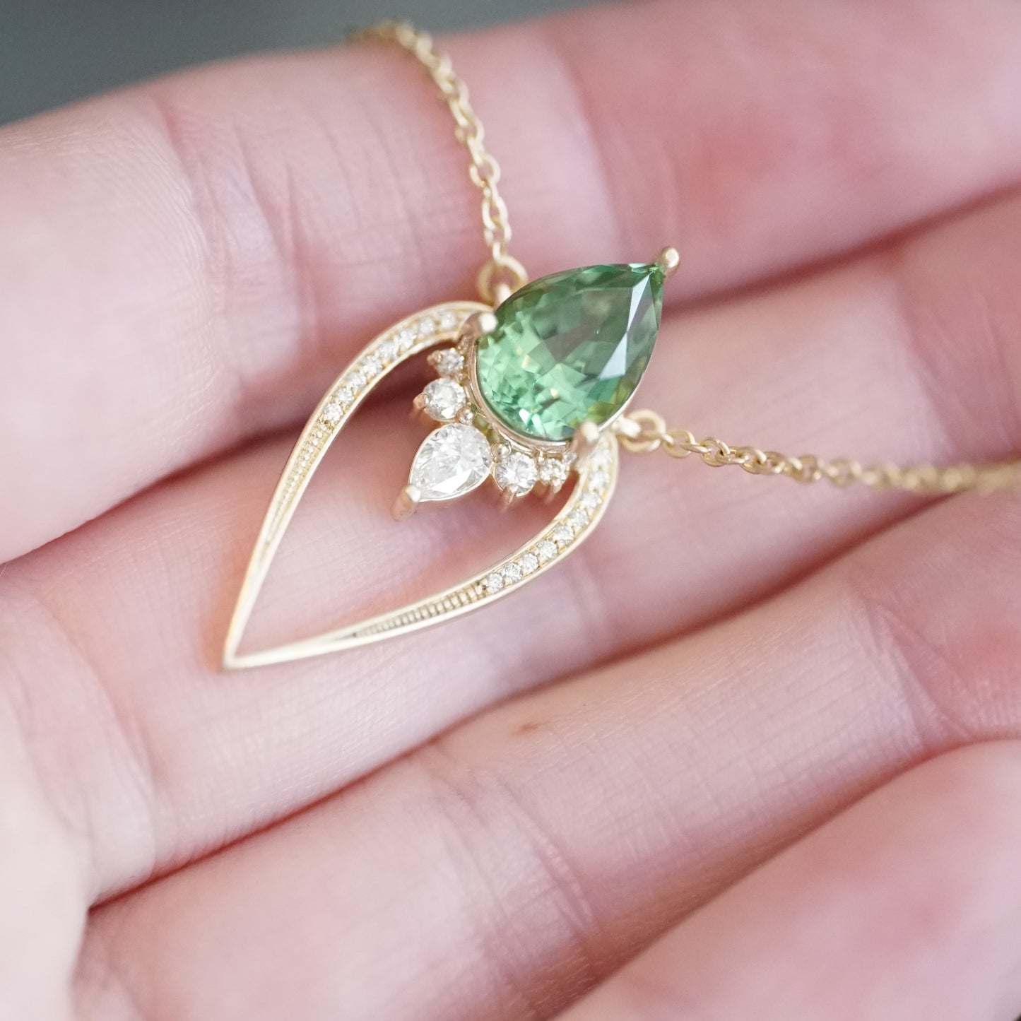 a yellow gold pendant with a vibrant green tourmaline pear shape and diamond accents suspended on a cable chain and laying on a light skinned left hand