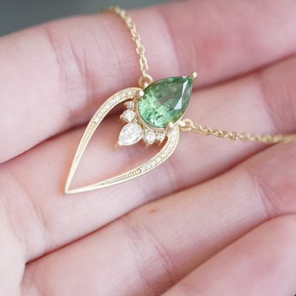 a yellow gold pendant with a vibrant green tourmaline pear shape and diamond accents suspended on a cable chain and laying on a light skinned left hand