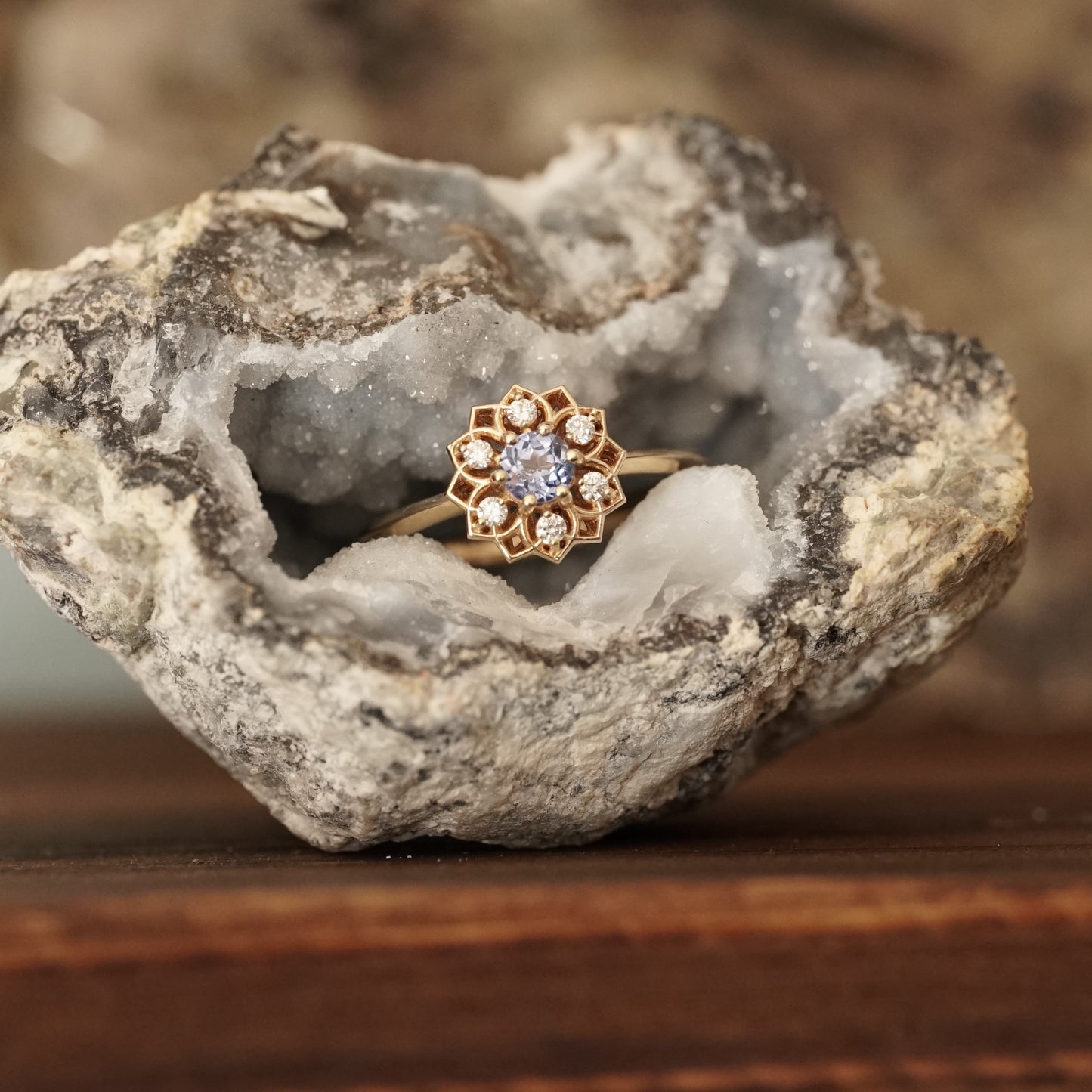 a yellow gold ring with a light blue sapphire enter stone and 6 white diamond accents in an open wire floral design lays in a sparkly geode on a wood base