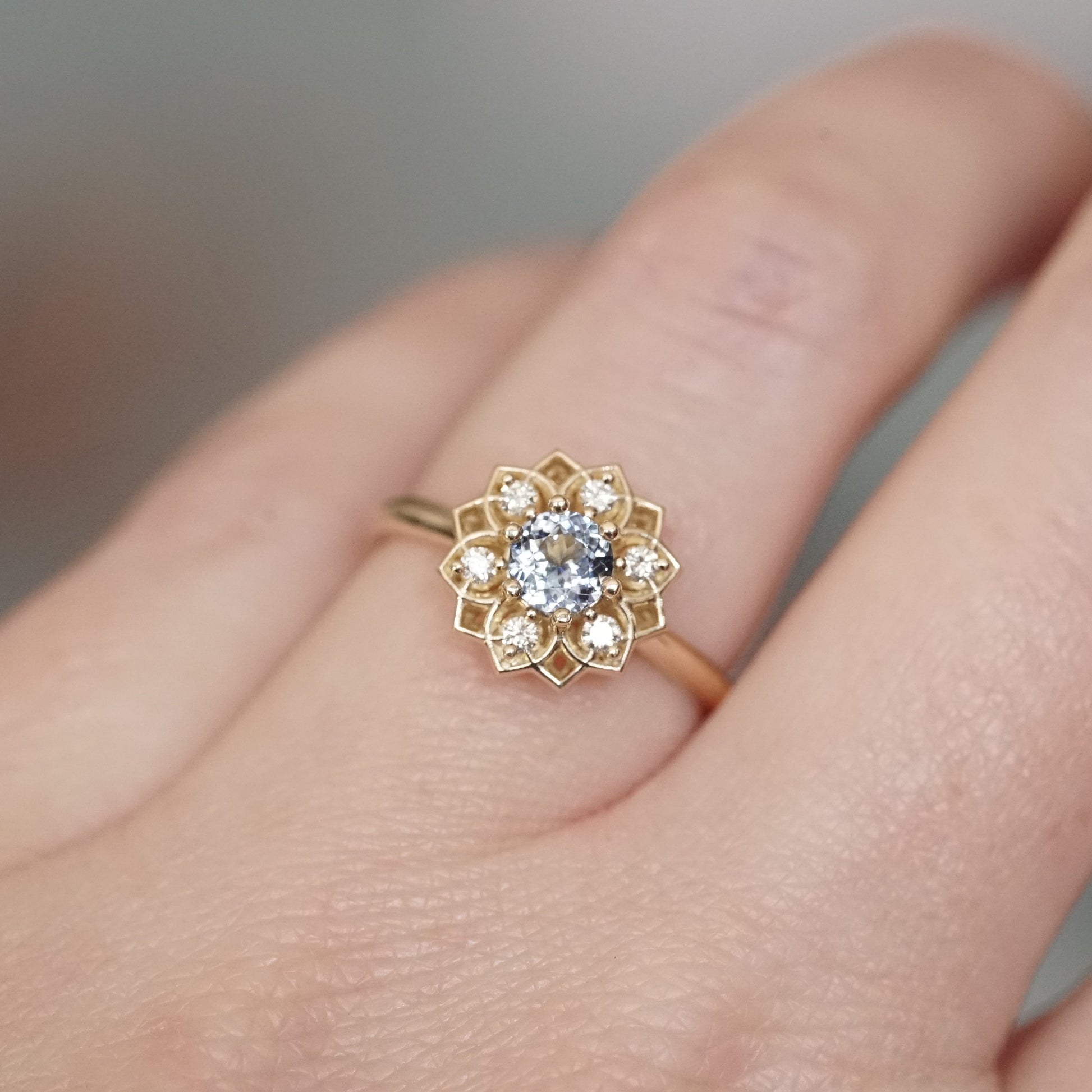 a yellow gold ring with a light blue sapphire enter stone and 6 white diamond accents in an open wire floral design on a light skinned left hand