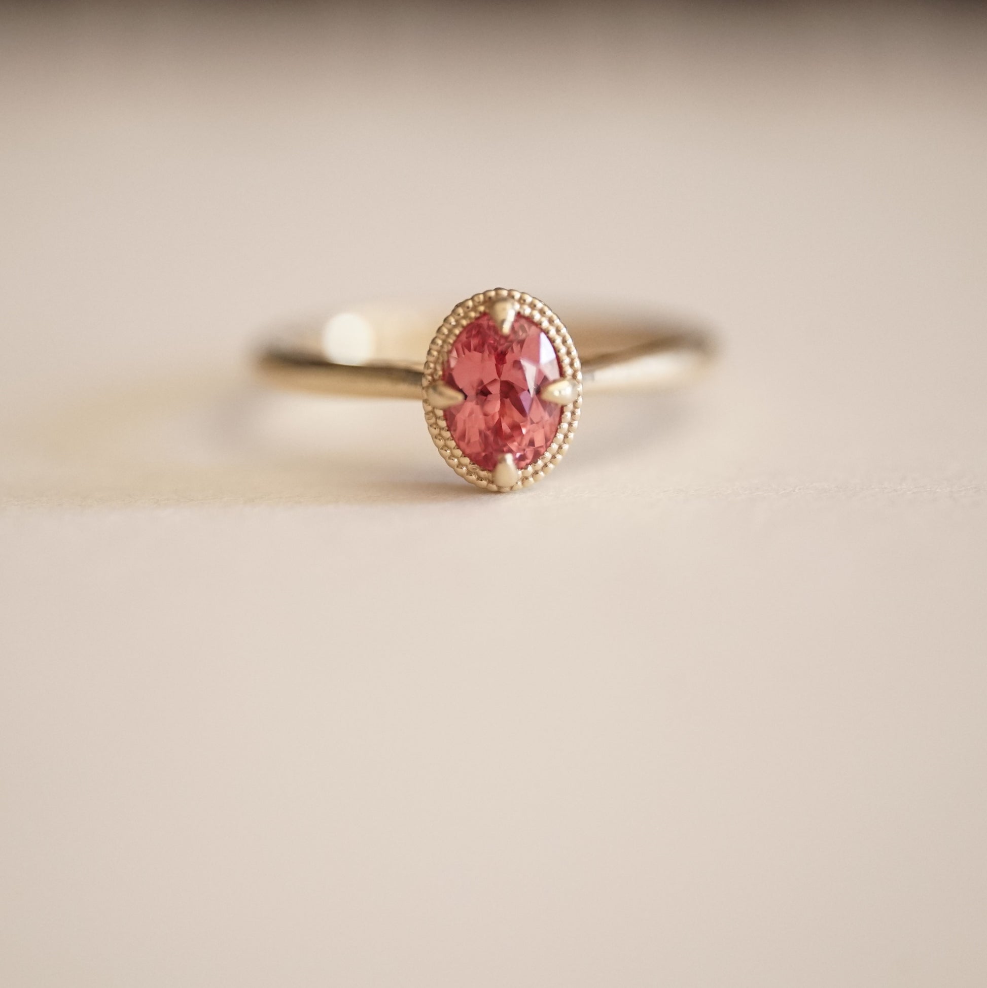 a macro photo of a vibrant oval pink spinel in a yellow gold ring setting with a beaded halo on a matte white background