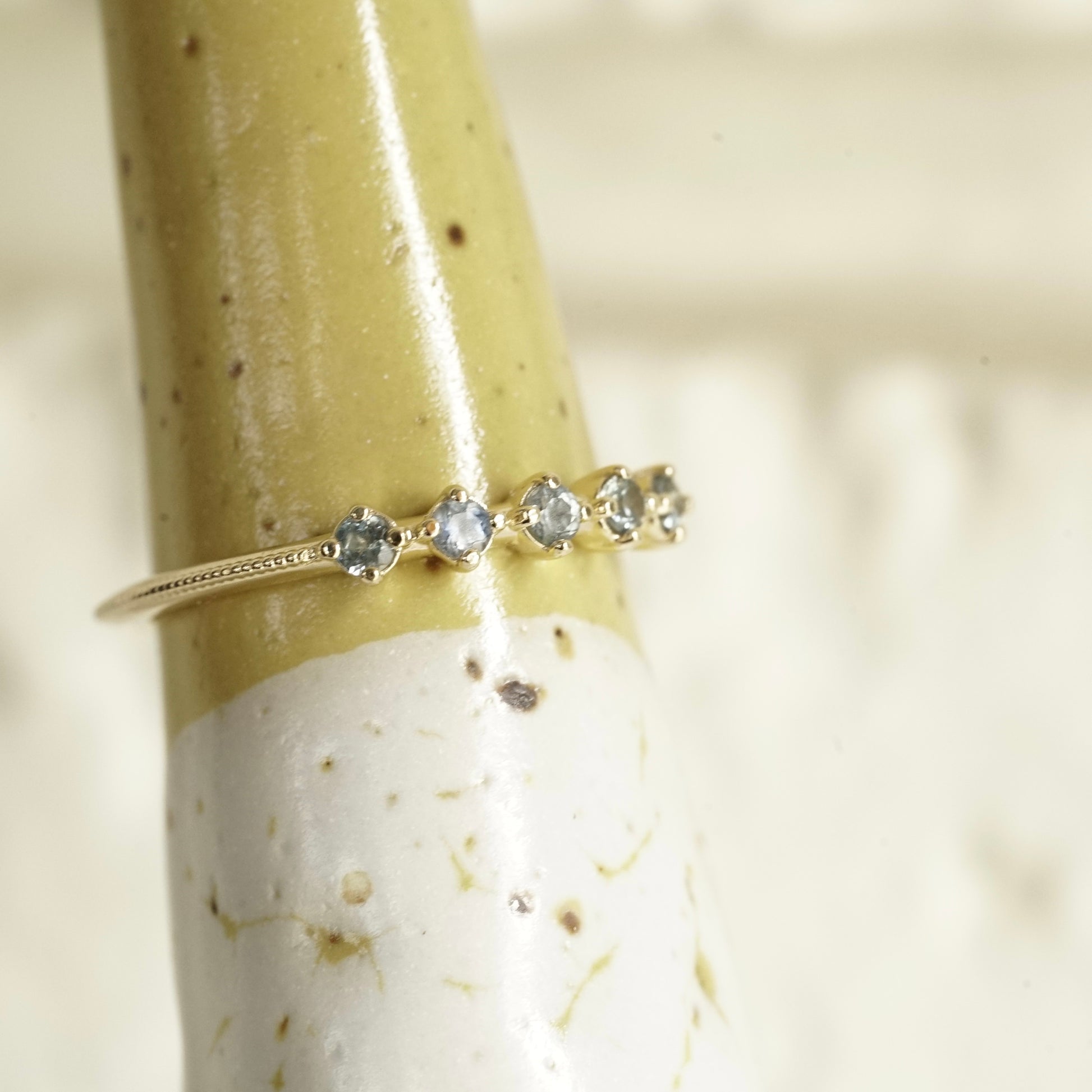a ring with 5 prong set light blue sapphires with migraine detailing on the shank on a yellow and white ceramic display