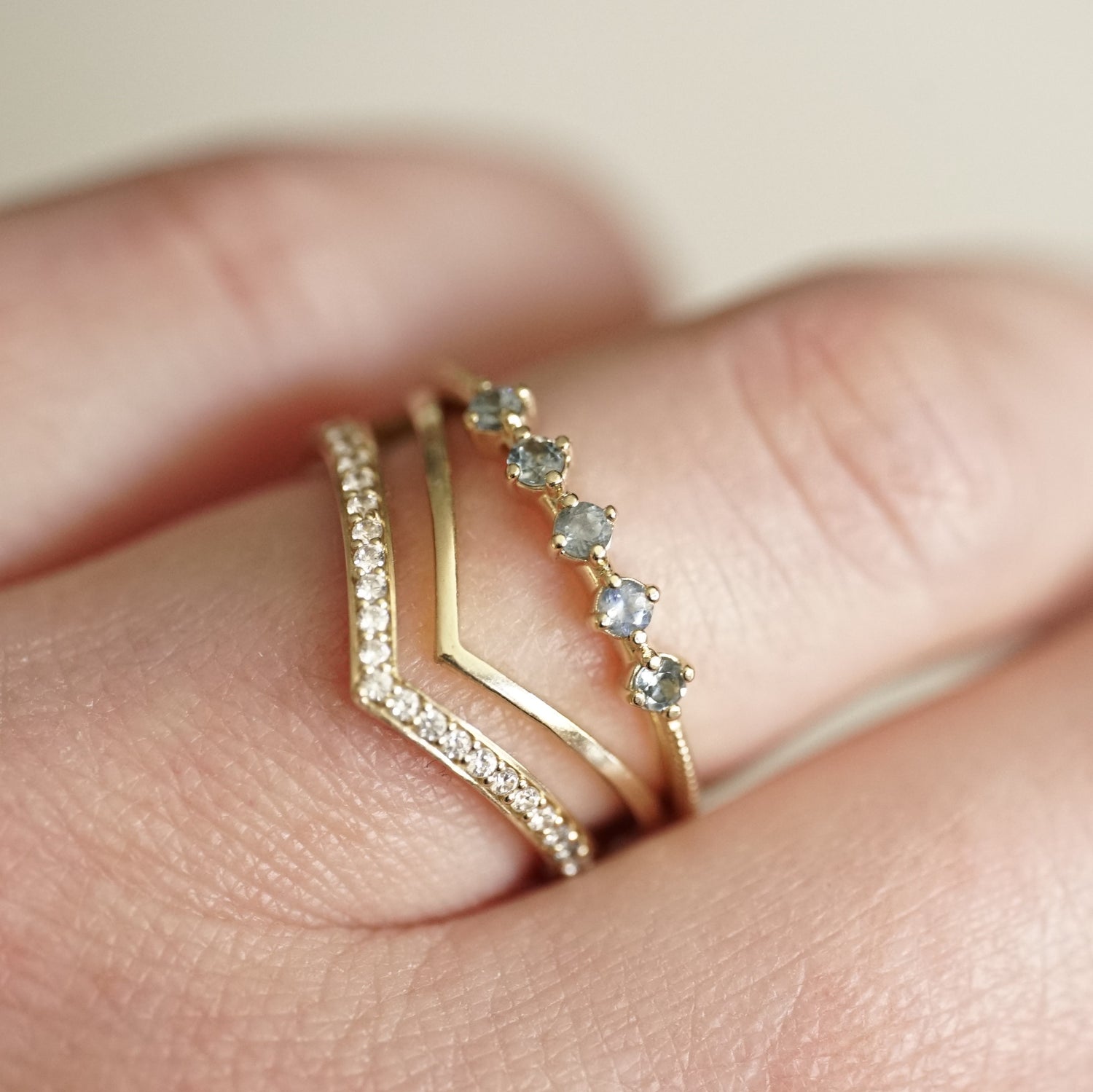a ring with 5 light blue sapphires in prong settings is stacked with 2 V shaped shadow stacking bands, with diamonds and one without on a light skinned left hand