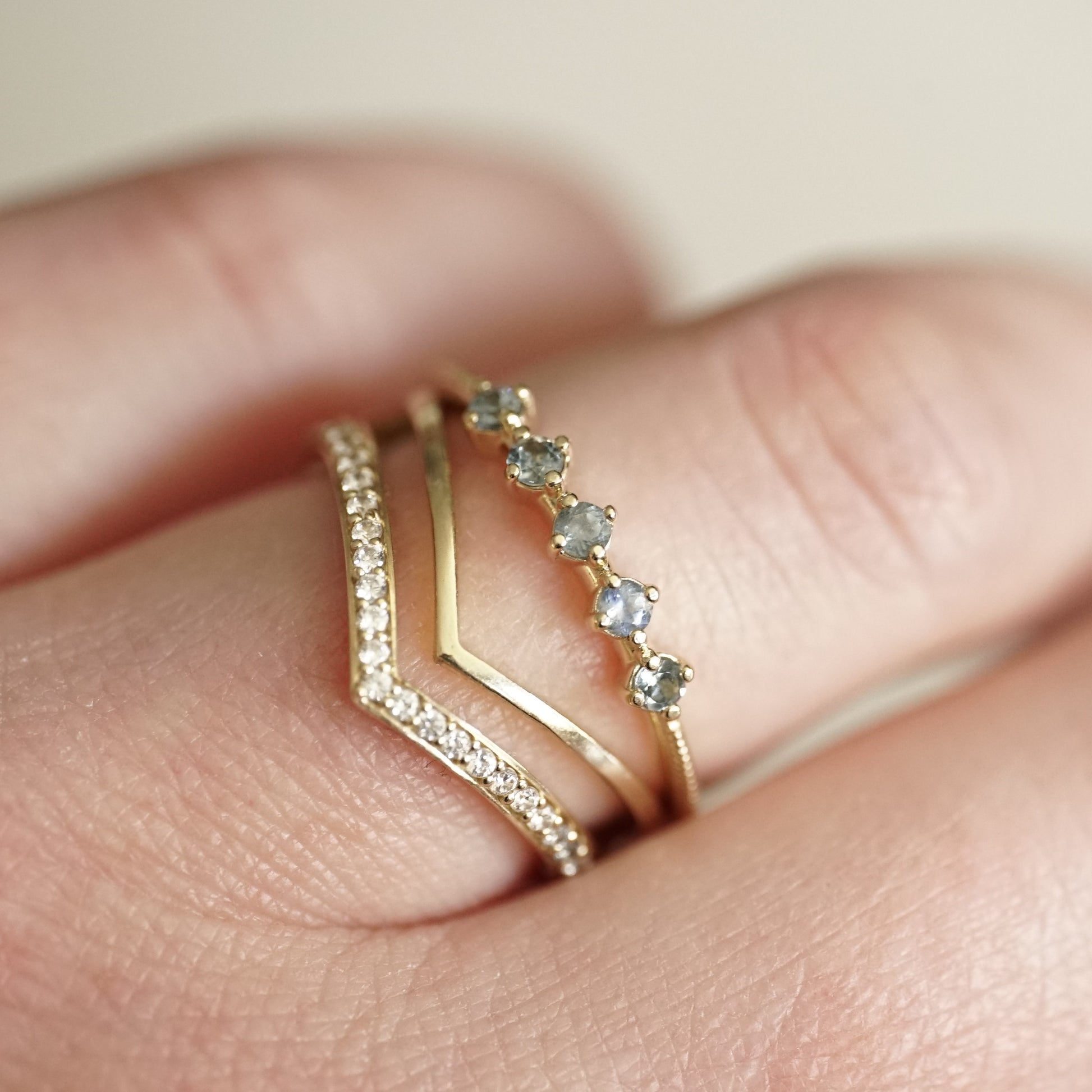 a ring with 5 light blue sapphires in prong settings is stacked with 2 V shaped shadow stacking bands, with diamonds and one without on a light skinned left hand