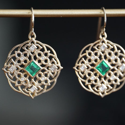 a pair of yellow gold earrings with princess cut emerald center stones and round and princess cut diamond earrings hang on a brass bar