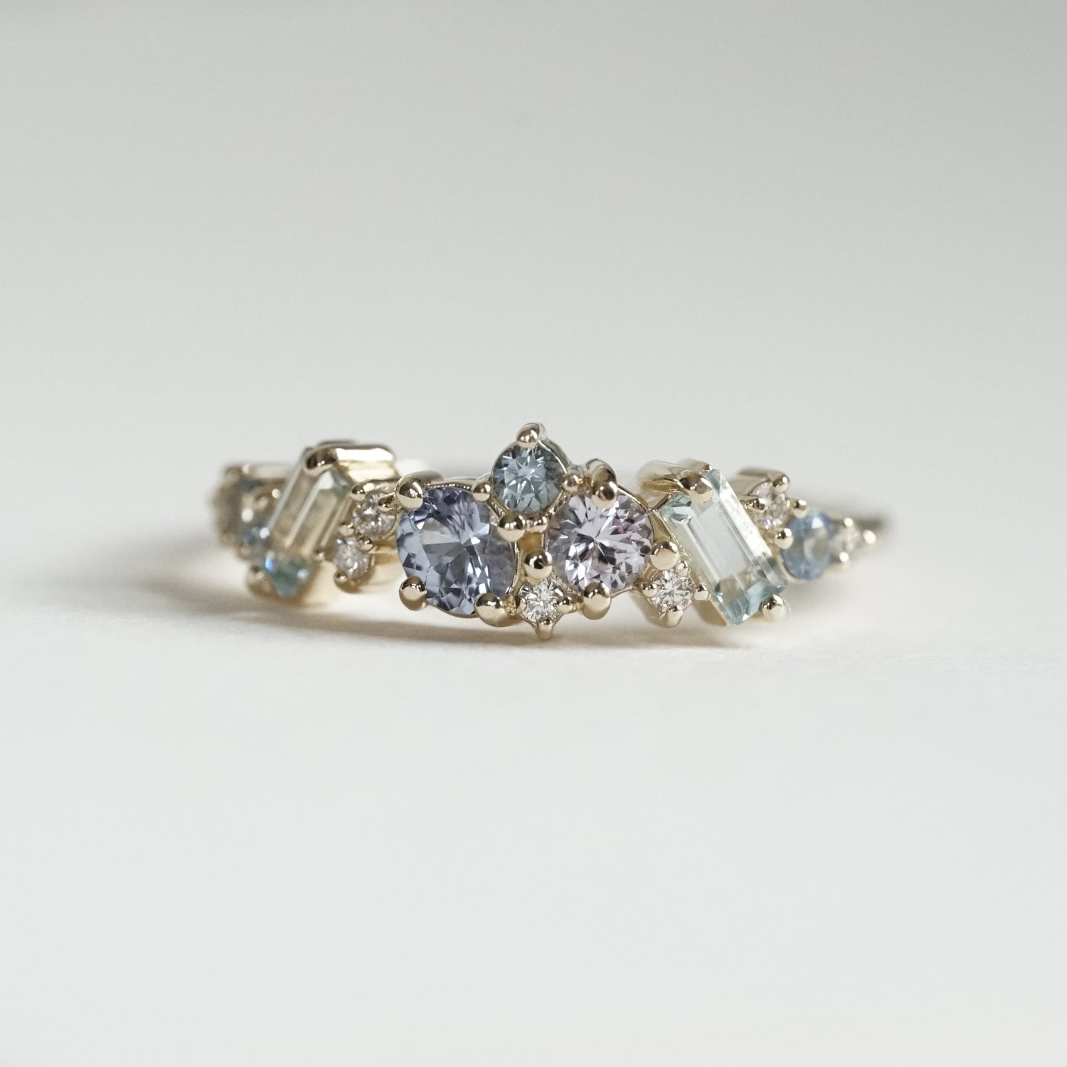 a macro photo of a yellow gold ring with blue gems of various colors sizes and shapes are mixed with round white diamonds on a matte white background