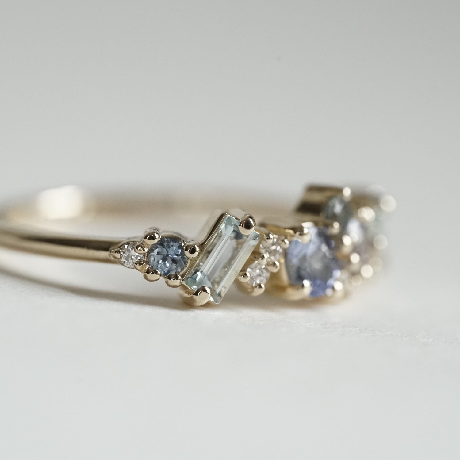 a macro photo of the side of a yellow gold ring with blue gems of various colors sizes and shapes are mixed with round white diamonds on a matte white background