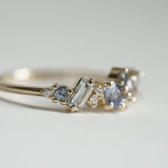 a macro photo of the side of a yellow gold ring with blue gems of various colors sizes and shapes are mixed with round white diamonds on a matte white background