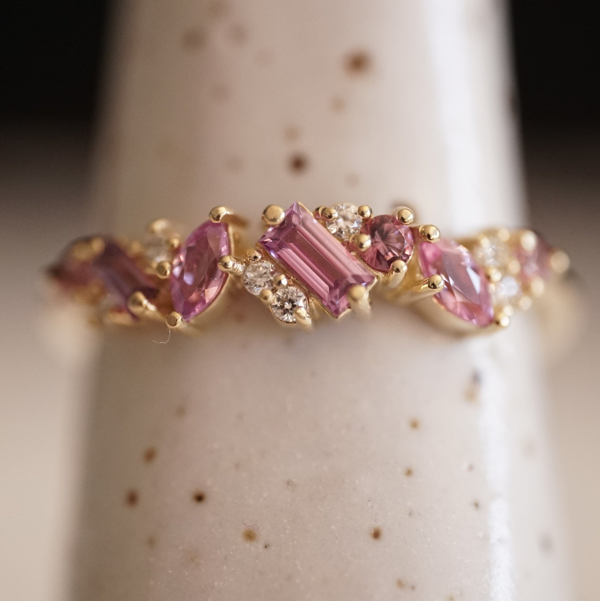 a macro photo of a yellow gold ring with pink sapphires of mixed shapes are paired with round white diamonds in a playful design on a white ceramic display