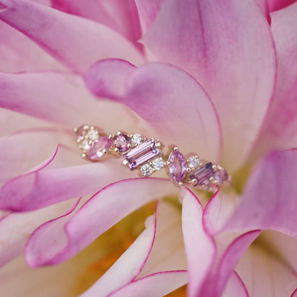 a yellow gold ring with pink sapphires of mixed shapes are paired with round white diamonds in a playful design. the ring is resting in a pink flower.