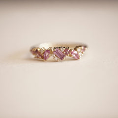 a yellow gold ring with pink sapphires of mixed shapes are paired with round white diamonds in a playful design rests on a matte white background